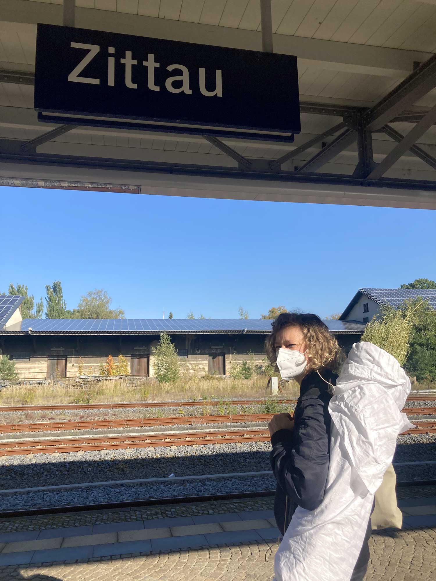 Ein Schild mit der Aufschrift Zittau am Bahnhof, darunter eine Frau mit Maske und einem Bündel auf dem Rücken